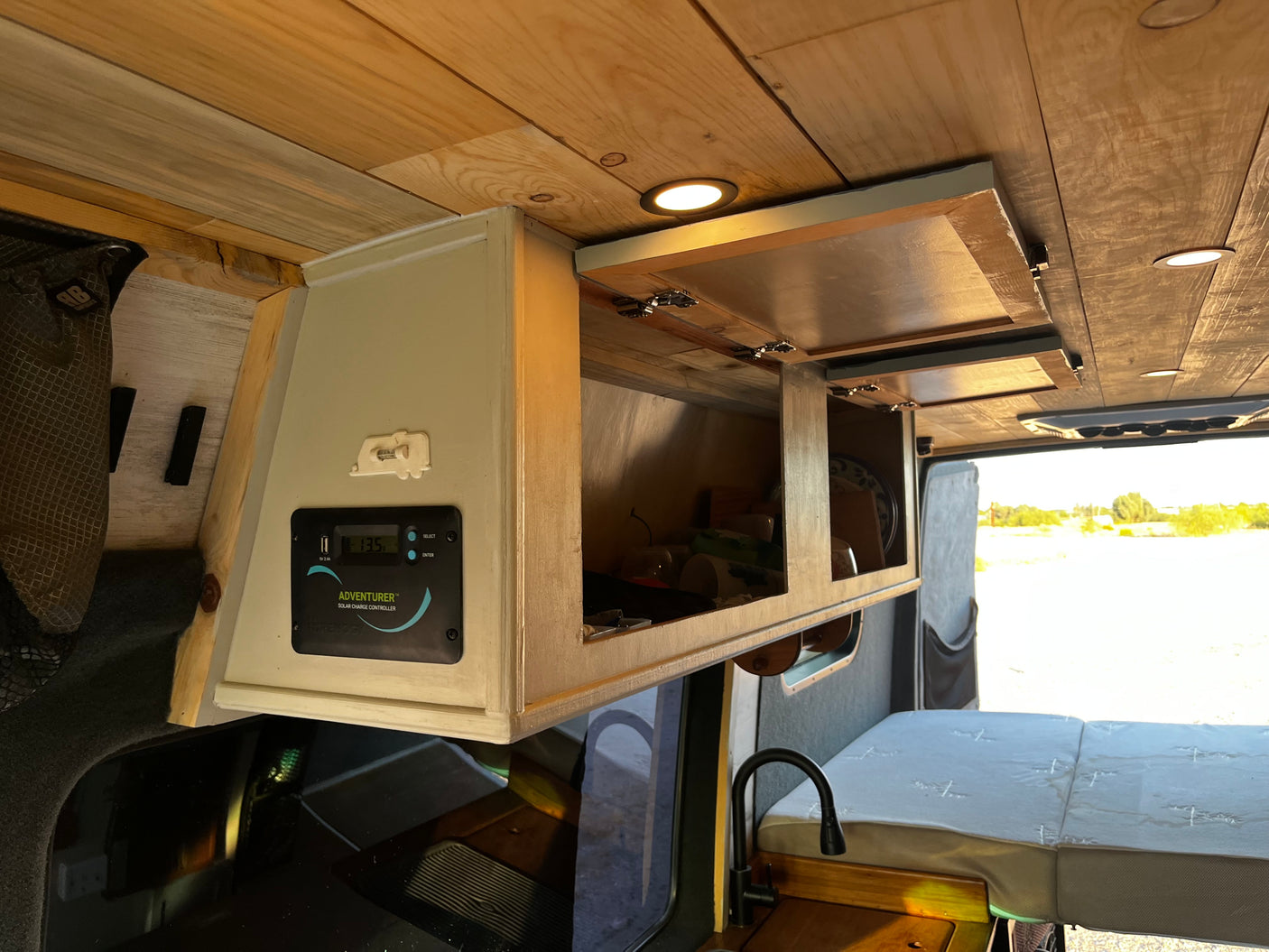 RV rental interior: custom wood ceiling & cabinets, solar charge controller, compact kitchen faucet, & light grey bed cushion.