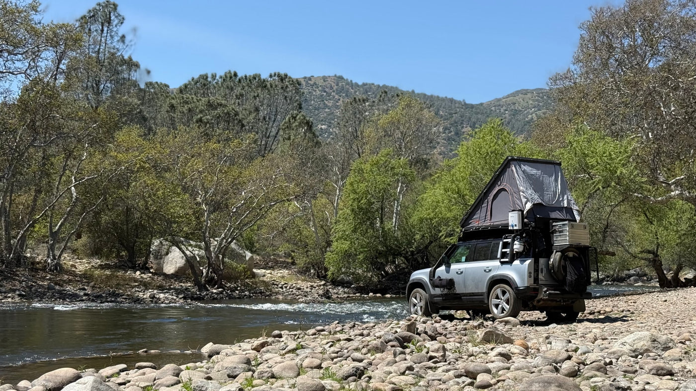 Overland-equipped SUV with rooftop tent on a rocky riverbank, surrounded by trees and mountains. An ideal RV rental for nature adventures. — photo 5