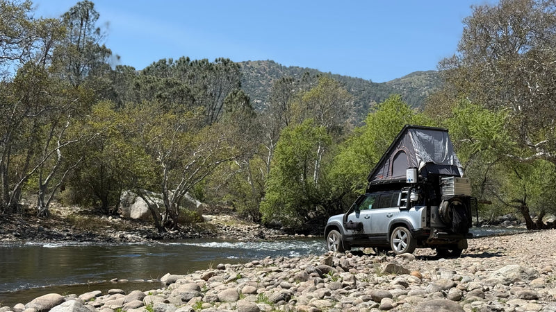 Overland-equipped SUV with rooftop tent on a rocky riverbank, surrounded by trees and mountains. An ideal RV rental for nature adventures. — photo 5