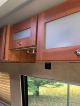 Inside an RV rental: overhead wood cabinets with frosted glass doors, and a window revealing trees and grass.