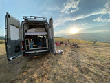 Camper van, rear doors open, shows bed & storage. A family relaxes on a golden hillside with mountain sunset views. Your ideal RV rental escape.