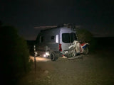 An RV rental with a dirt bike on its rear rack, parked on a dark, unpaved road at night for off-grid adventure camping.