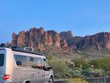Silver 'Serenity' RV rental camper van parked by desert plants, facing majestic, rugged mountains under a clear sky.