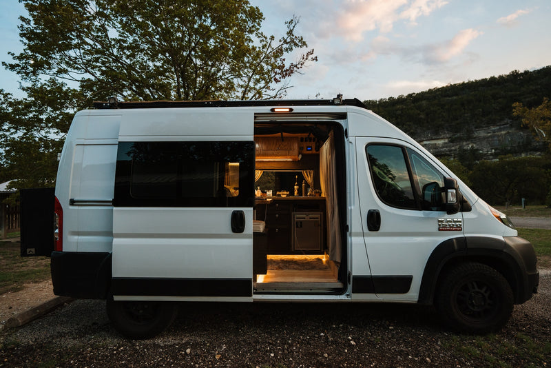 Step inside this cozy Ram ProMaster RV rental, featuring a custom interior kitchen with warm lighting, parked amidst nature. — photo 3