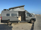 An rv rental gray camper van with a pop-top roof, open side door revealing its interior, and rooftop AC unit, parked by the road.