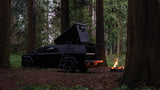 This RV rental is a black Tesla Cybertruck with a pop-up tent, perfect for camping in the forest by a campfire.