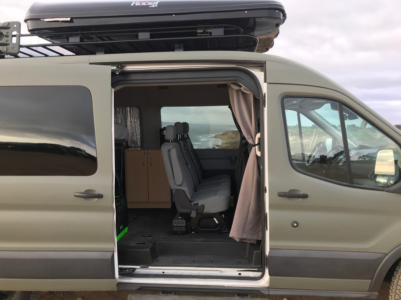 This spacious rv rental camper van with an open side door displays its interior seating and storage, topped with a cargo box, overlooking a scenic coast. — photo 14