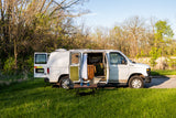 Converted white van, an ideal RV rental, parked in a sunny grassy spot. Doors open to show interior; chairs & table outside.