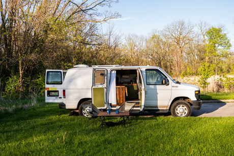 Converted white van, an ideal RV rental, parked in a sunny grassy spot. Doors open to show interior; chairs & table outside.