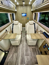 Stylish RV rental interior with two diamond-stitched captain's chairs, fold-out tables, a rear TV, and wood flooring.