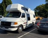 White Class B rv rental parked in a sunny lot next to a grey Honda Civic. Trees visible in the background.