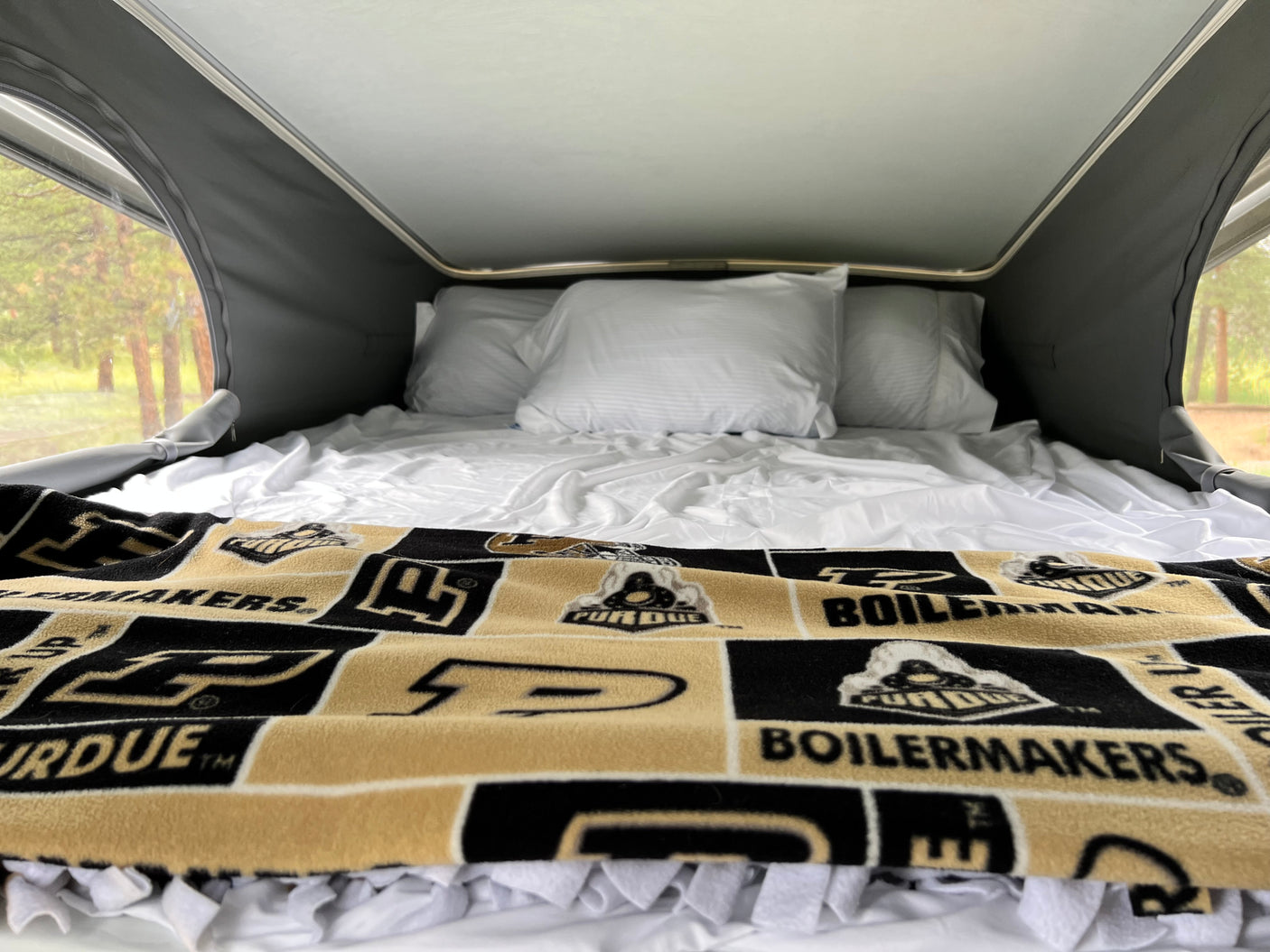 Inside an RV rental pop-top: a comfy bed with white linens and a Purdue Boilermakers blanket, surrounded by nature views.