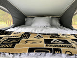 Inside an RV rental pop-top: a comfy bed with white linens and a Purdue Boilermakers blanket, surrounded by nature views.