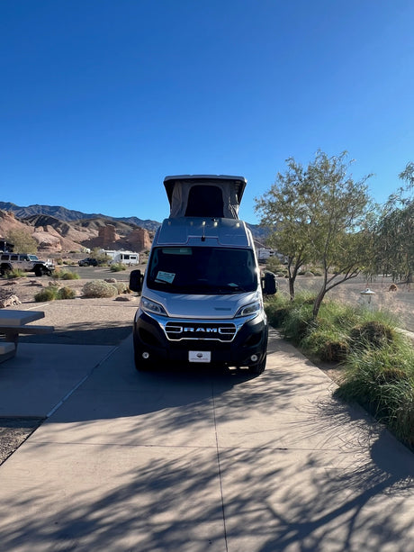 Front view of a silver RV rental with a pop-top at a scenic desert campsite. Mountains and clear blue skies abound.