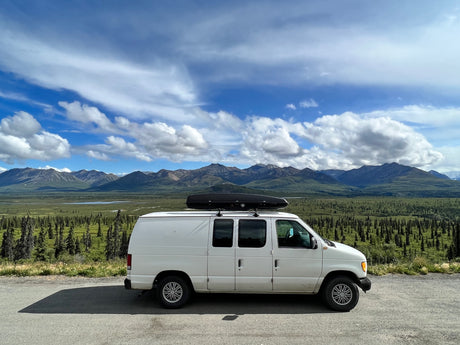 White RV rental van overlooks stunning mountains, a forest, and lakes under a blue sky with white clouds.
