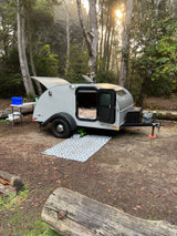 Teardrop RV rental set up in a misty forest campsite at sunrise, with a cozy bed visible inside.