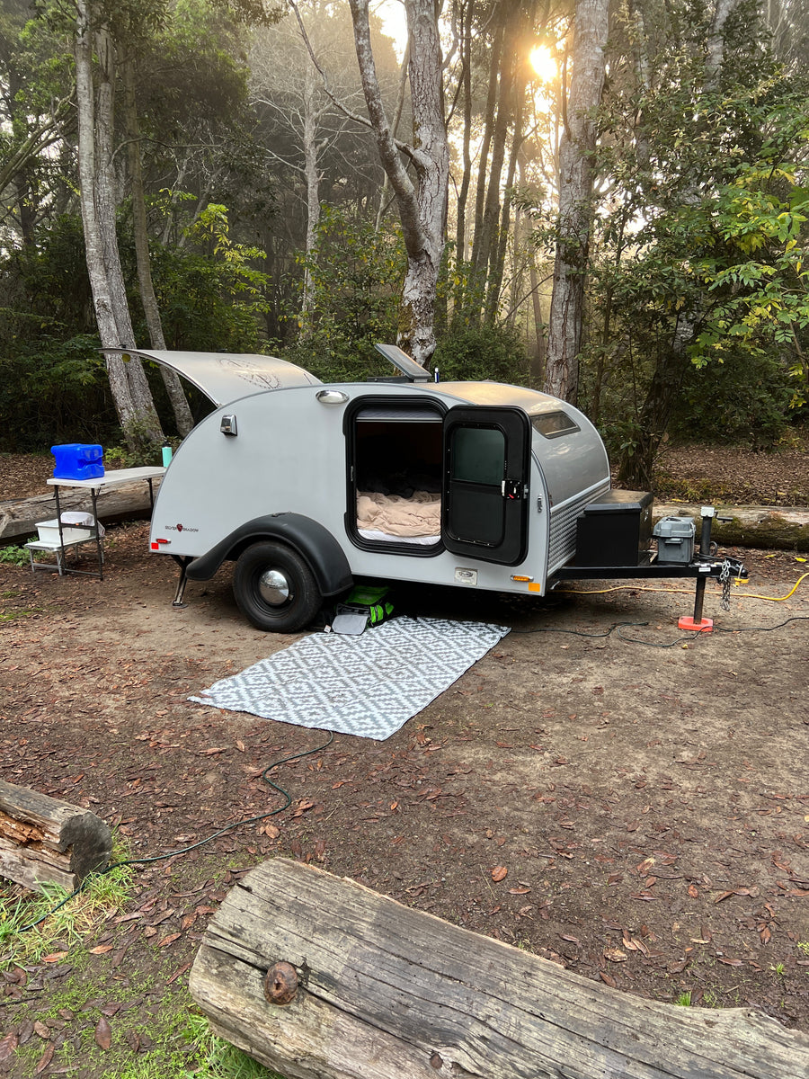 Teardrop RV rental set up in a misty forest campsite at sunrise, with a cozy bed visible inside.