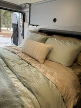 Relax in this cozy RV rental bed, featuring soft green and beige linens with a winter forest view.