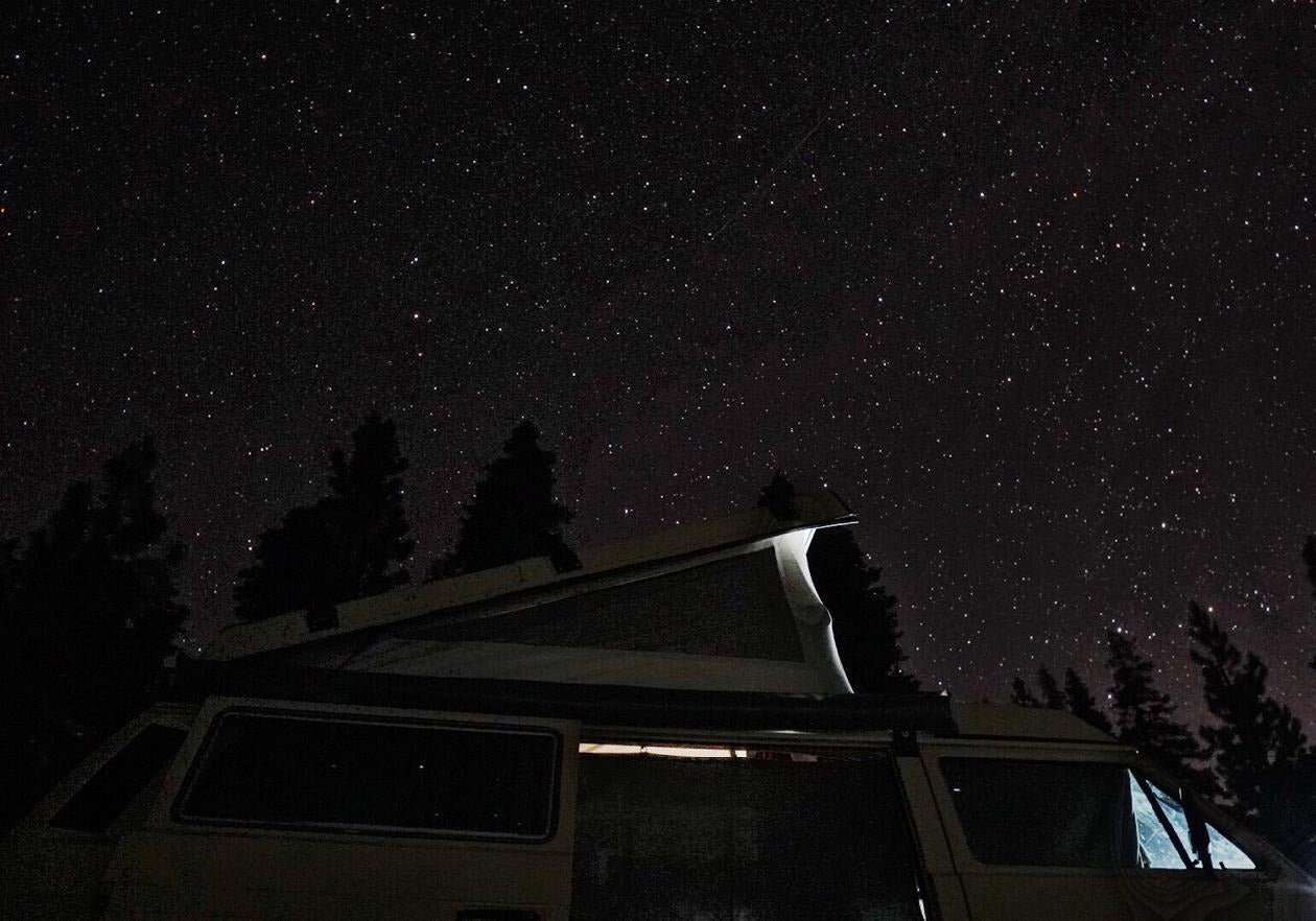 An RV rental, a camper van with a pop-top, parked under a brilliant, star-filled night sky with silhouetted trees.