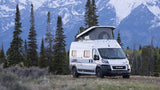 White camper van RV rental parked in a beautiful mountain landscape with tall pine trees and a grassy field.