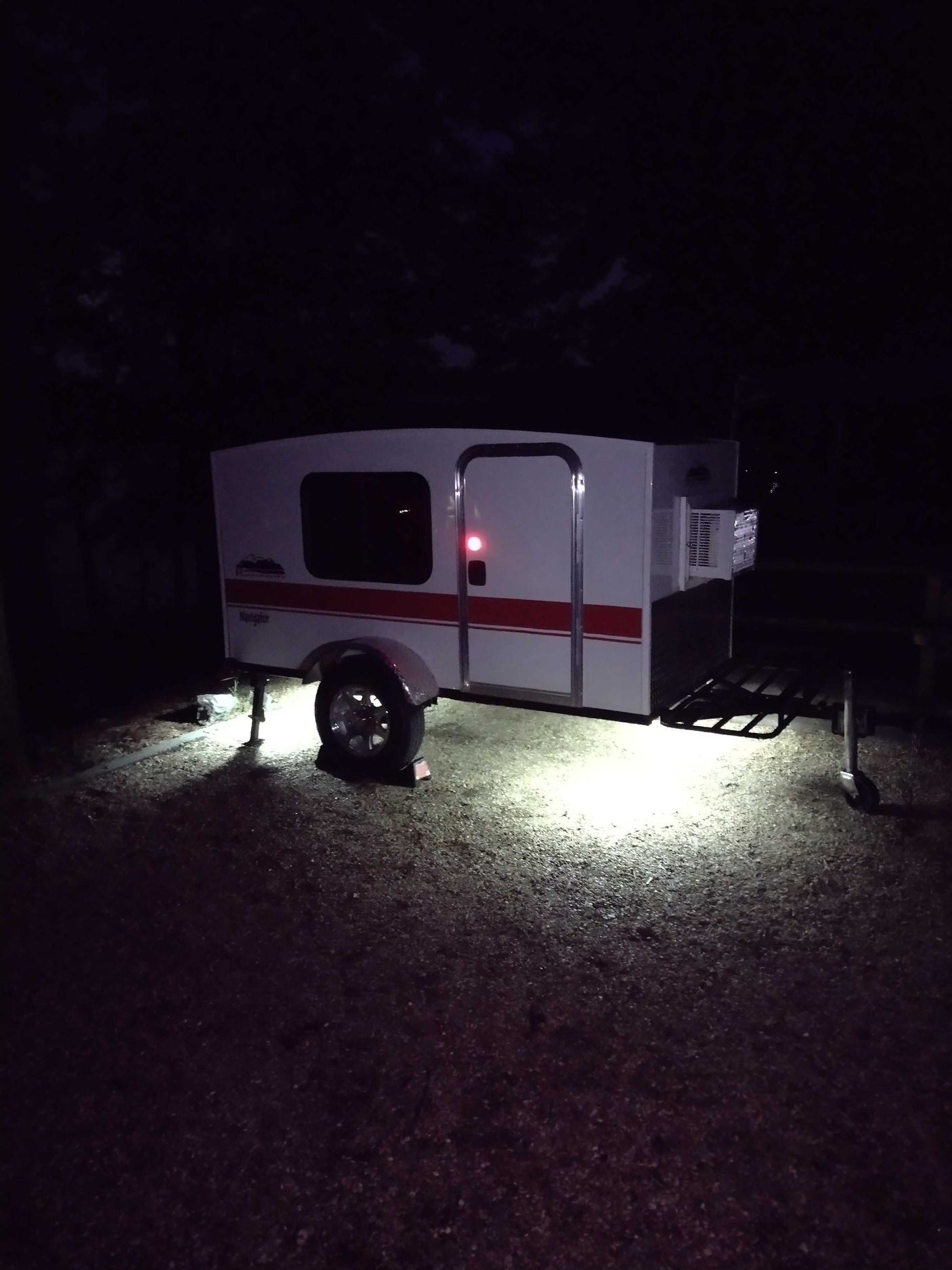White teardrop camper RV rental lit by ground lights at night. Features include an AC unit and red stripe.