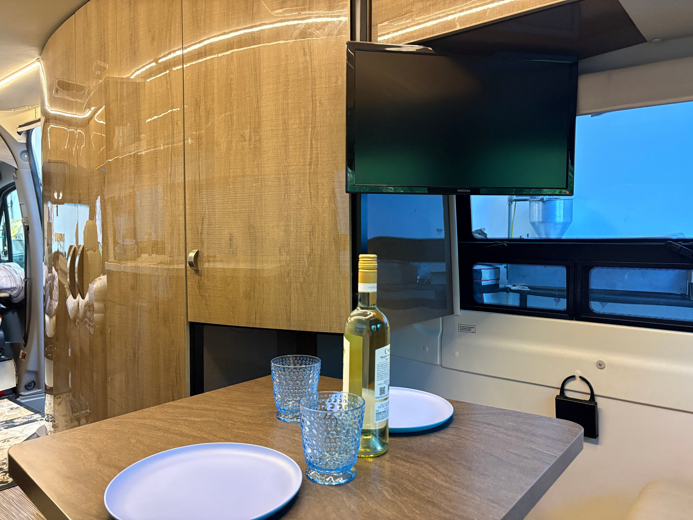 Inside an rv rental: a dining table with wine, glasses, and plates. A TV is mounted above. Light wood walls create a cozy atmosphere.