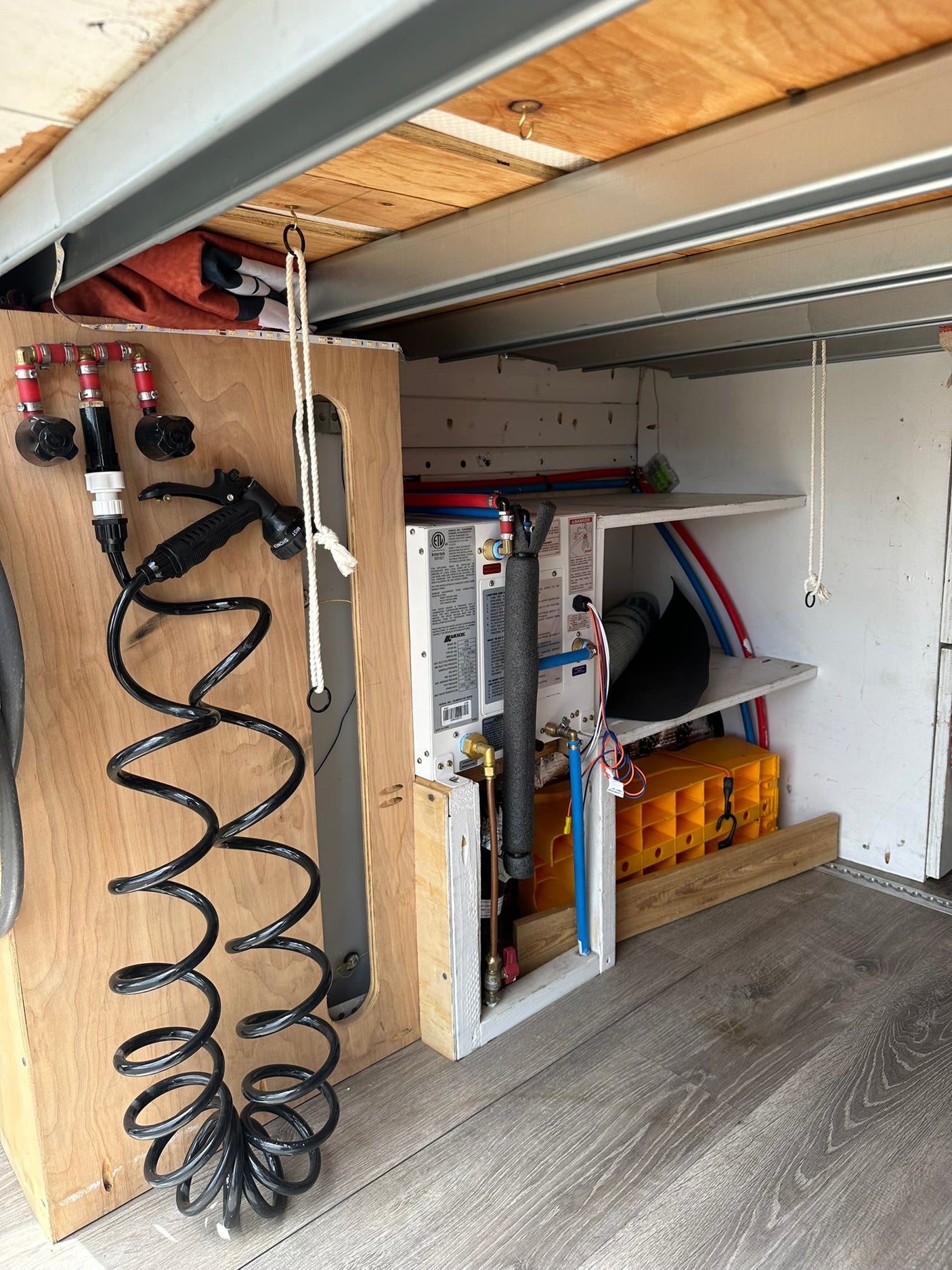 An RV rental utility area with an on-demand water heater, organized plumbing, and a coiled shower hose. Efficient camp system.