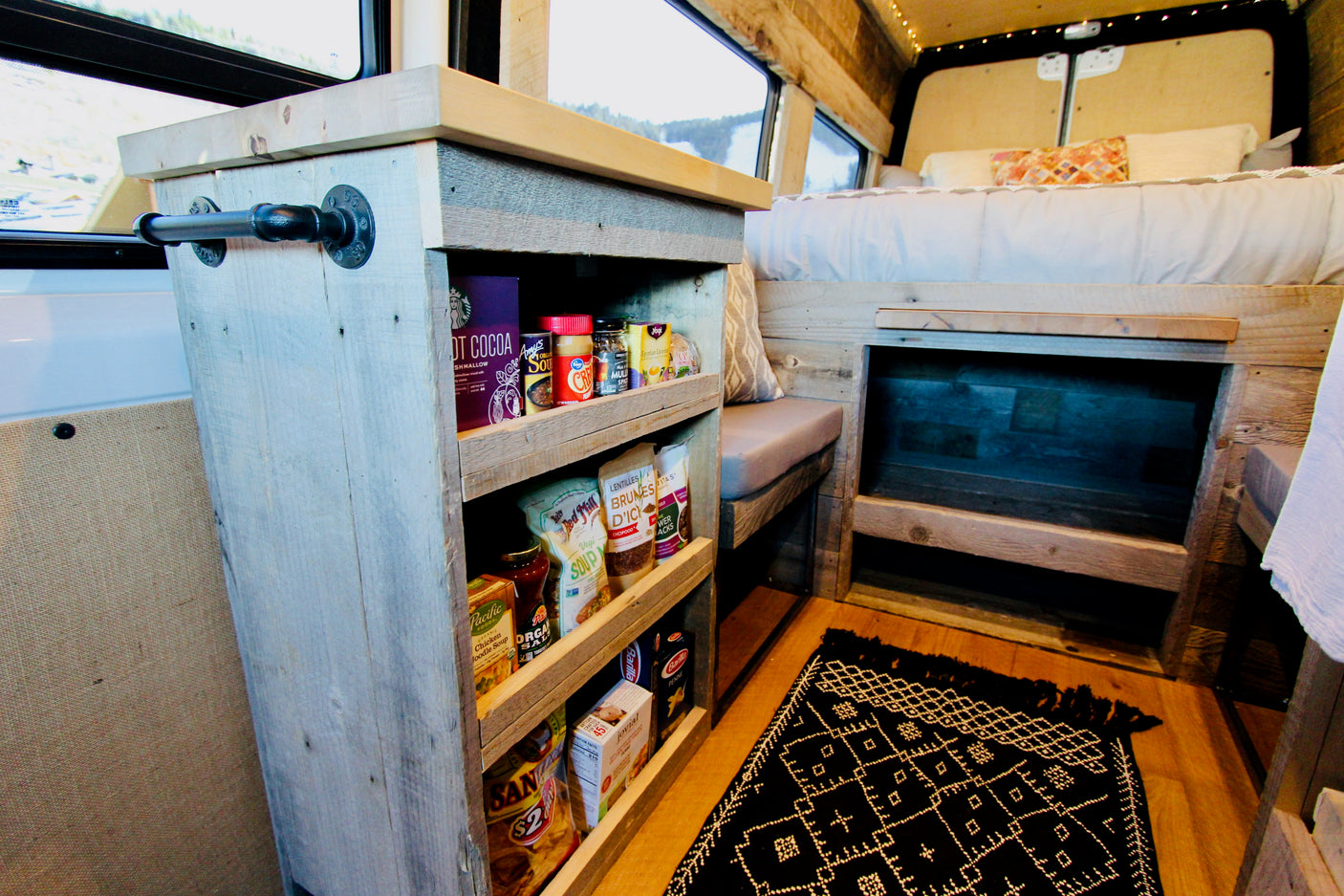 RV rental interior: Built-in wood shelves stocked with food, a cozy bed, and a patterned rug for comfortable mobile living.