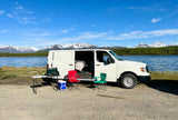 White RV rental van parked lakeside with majestic snow-capped mountains. Complete with camp setup: stove, chairs, cooler. Perfect for outdoor adventures.