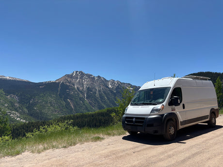 This RV rental, a white camper van, enjoys a scenic mountain view with snow-capped peaks and lush forests.