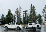 White truck towing a Viking outdoor RV rental trailer on wet asphalt, among tall pine trees. Perfect for your next adventure!