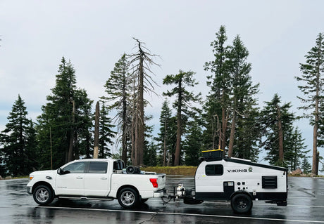 White truck towing a Viking outdoor RV rental trailer on wet asphalt, among tall pine trees. Perfect for your next adventure!