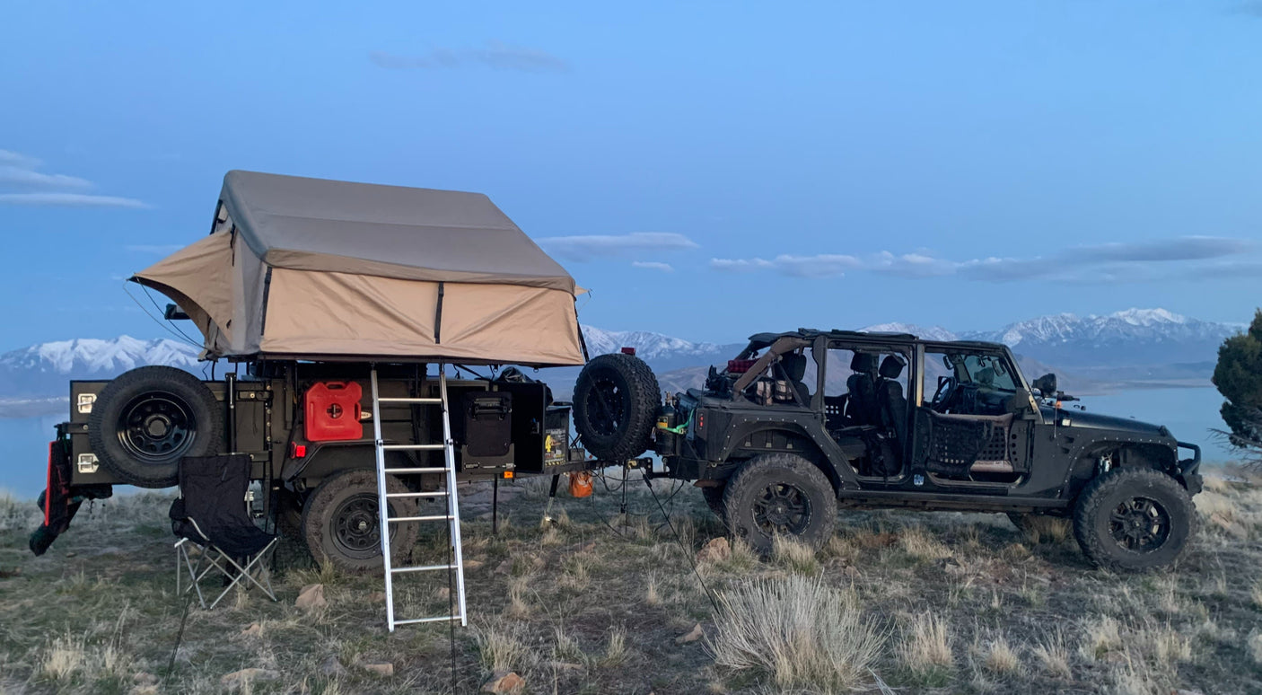 Overland RV rental setup: Jeep towing an off-road trailer with a rooftop tent, by a lake and snow-capped mountains.