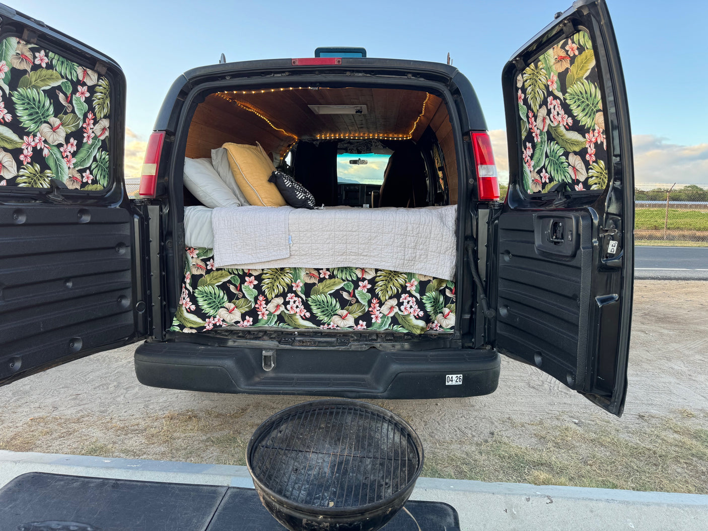 Inside an open **rv rental** camper van: bed with tropical decor, string lights, and grill setup for outdoor adventure.