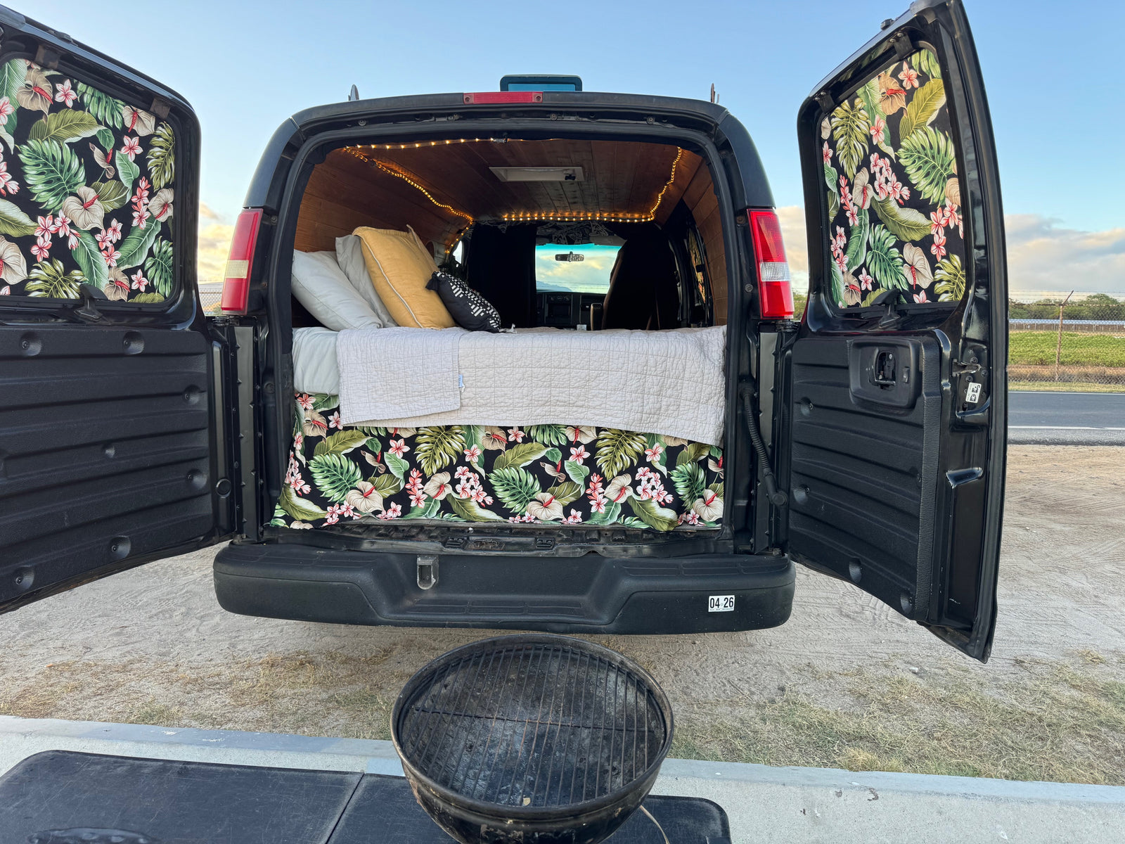 Inside an open **rv rental** camper van: bed with tropical decor, string lights, and grill setup for outdoor adventure.