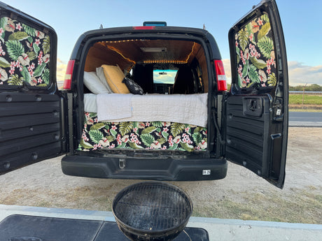 Inside an open **rv rental** camper van: bed with tropical decor, string lights, and grill setup for outdoor adventure.