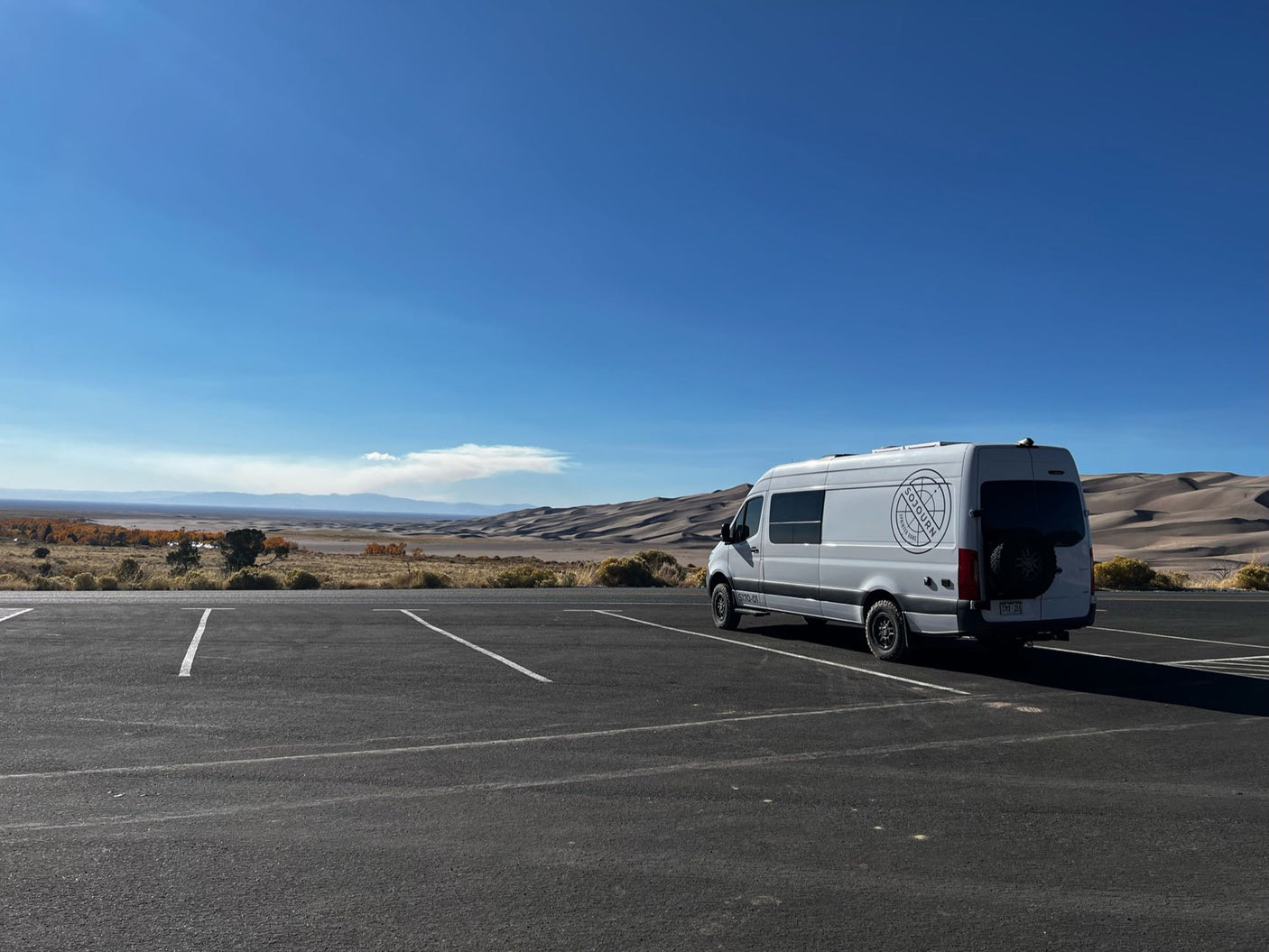 White RV rental camper van parked in front of majestic sand dunes & distant mountains under a clear blue sky. Ready for adventure!