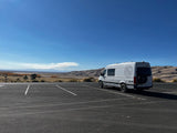 White RV rental camper van parked in front of majestic sand dunes & distant mountains under a clear blue sky. Ready for adventure!