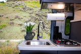 Inside an RV rental: a modern kitchen with sink, stovetop, and flowers, offering a scenic hillside view from the window.