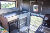 Compact RV rental interior: kitchen with fridge, sink, stove, storage, and a scenic window view.