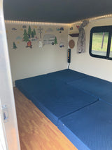RV rental interior: blue fold-out mattresses, wood-look floor, charming camping wall decals, and warm string lights.