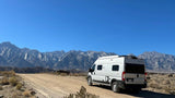 An RV rental sits on a dirt road, with a majestic mountain range under a clear blue sky in the background.