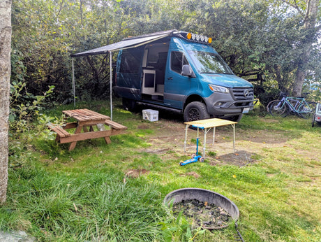 RV rental ready for adventure: A blue camper van with an awning, picnic table, and bikes at a wooded campsite.