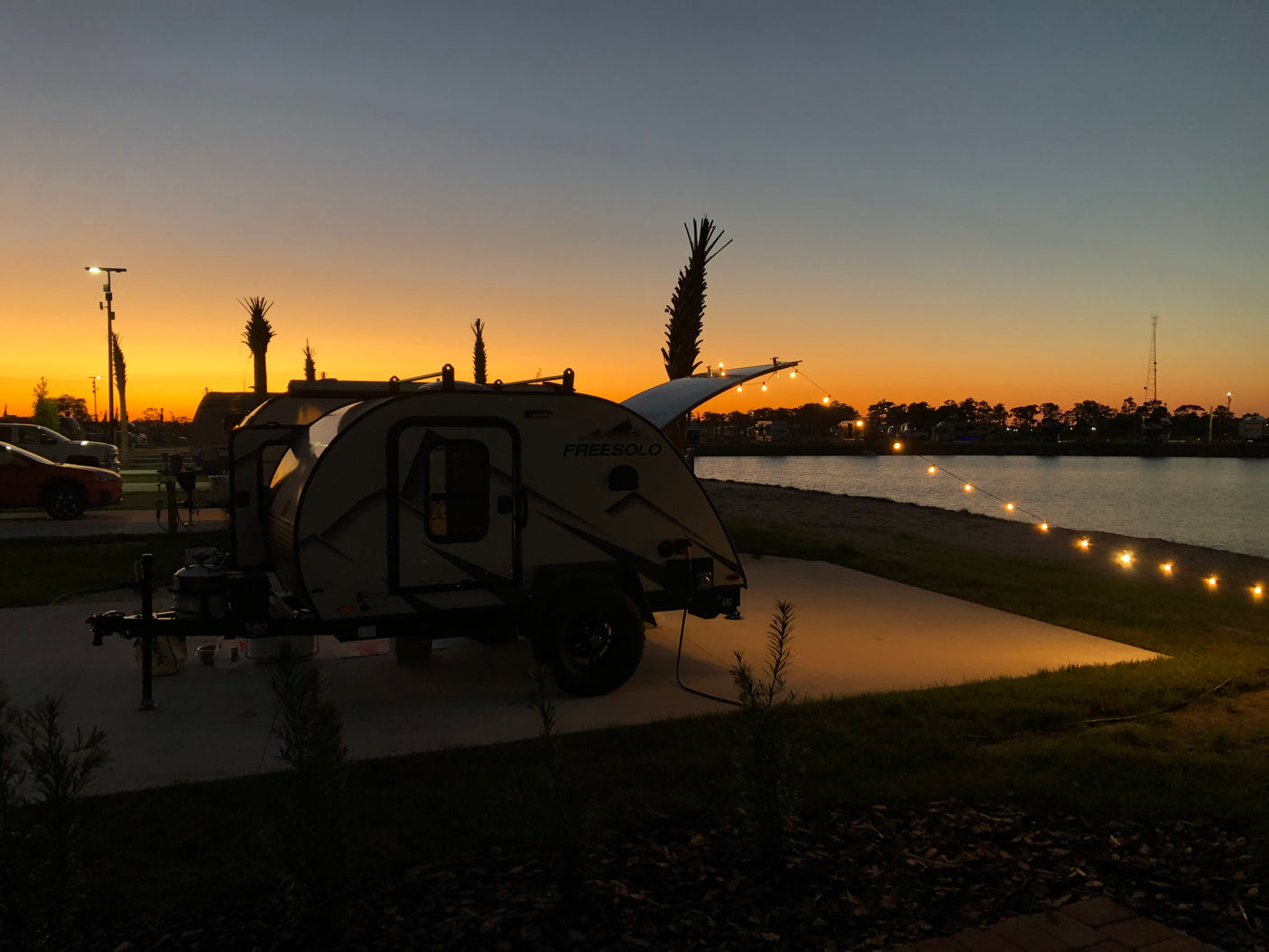 Enjoy a beautiful sunset at a waterfront RV rental campsite, featuring a 'Freesolo' teardrop trailer and string lights.