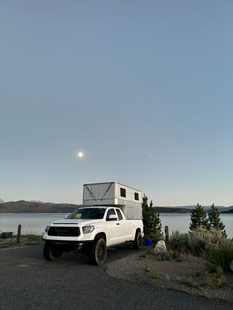 White Toyota Tundra with RV truck camper parked by a serene lake under a moonlit sky. Ideal for an rv rental adventure!