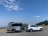 White SUV towing a silver travel trailer (similar to an RV rental) with scenic mountains and trees.