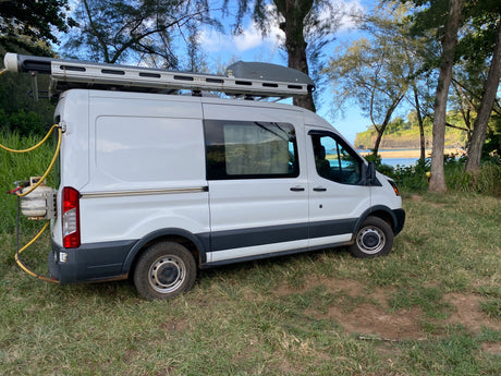 White Ford Transit rv rental camper van parked on grass by a scenic beach, equipped with roof gear for outdoor living.