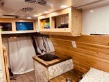 Bright rv rental interior, showing wooden walls, integrated sink/cooktop, overhead storage, and a sleeping area.