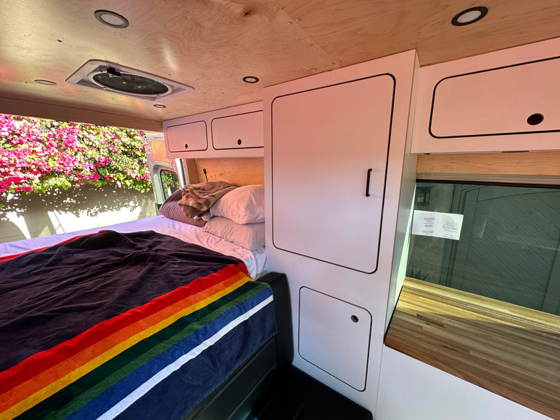 Cozy camper van interior for your next RV rental, featuring a bed, storage, and natural light from a window overlooking flowers. — photo 22