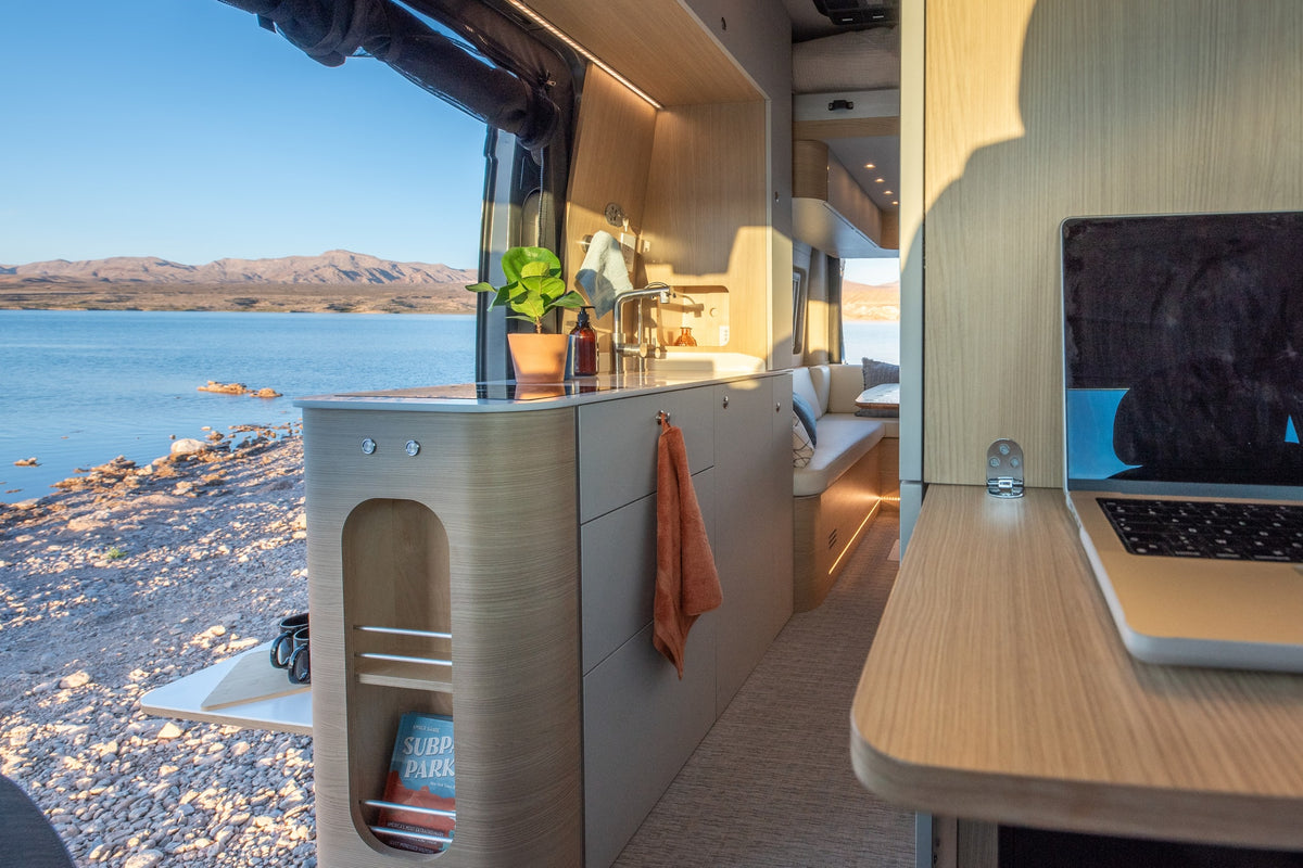 Modern RV rental interior with a laptop desk and kitchen, overlooking a serene lake and mountains. Perfect for van life! — photo 7